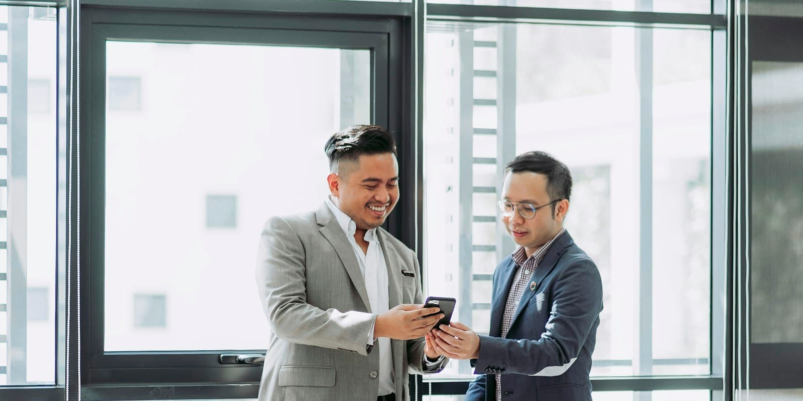 Two colleagues in suits looking at a phone together by an office window
