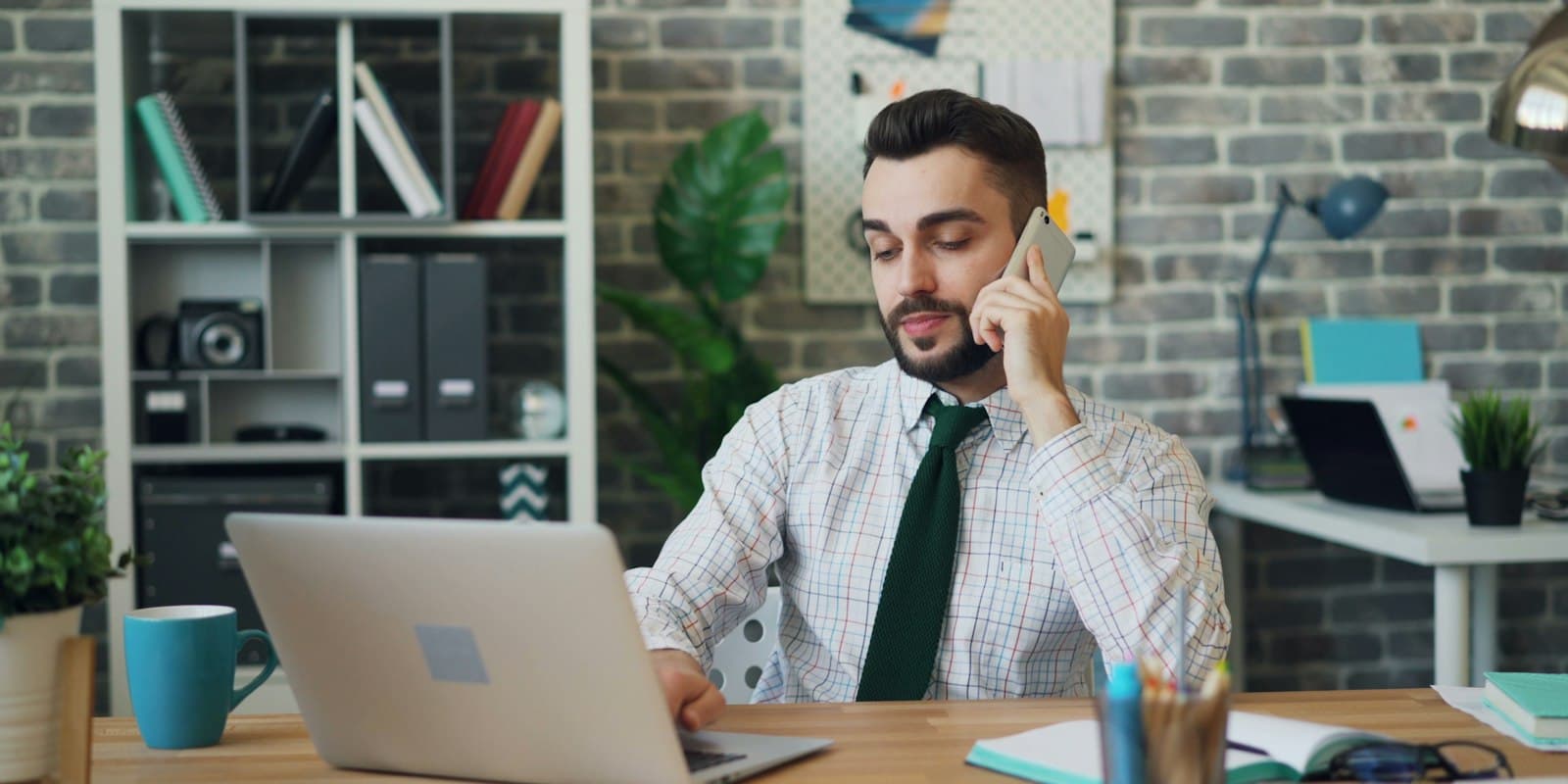 A man in a shirt and tie talking on his phone at a desk with a laptop