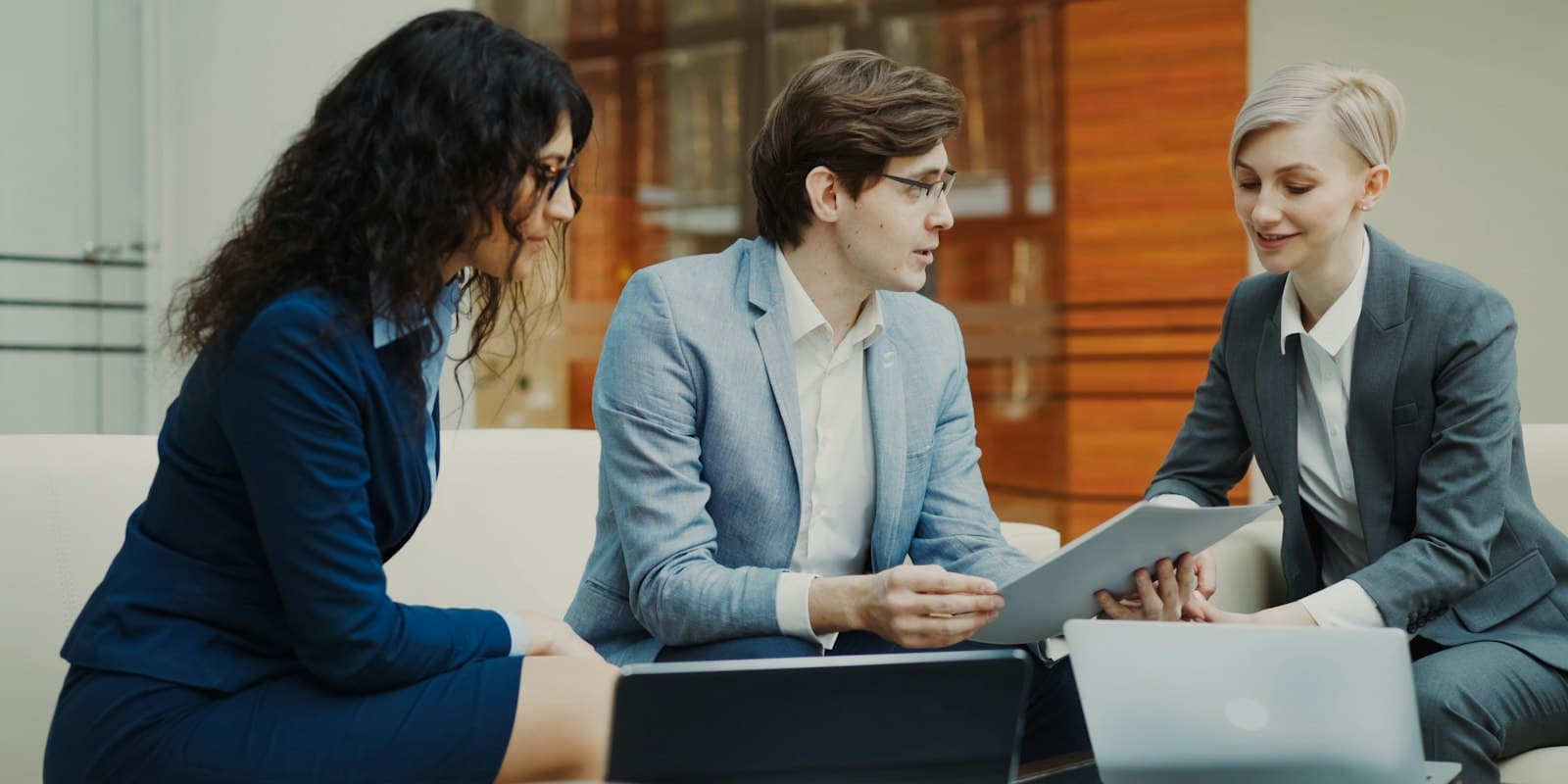Three colleagues on a sofa go over a document together, with two laptops open on the table in front of them