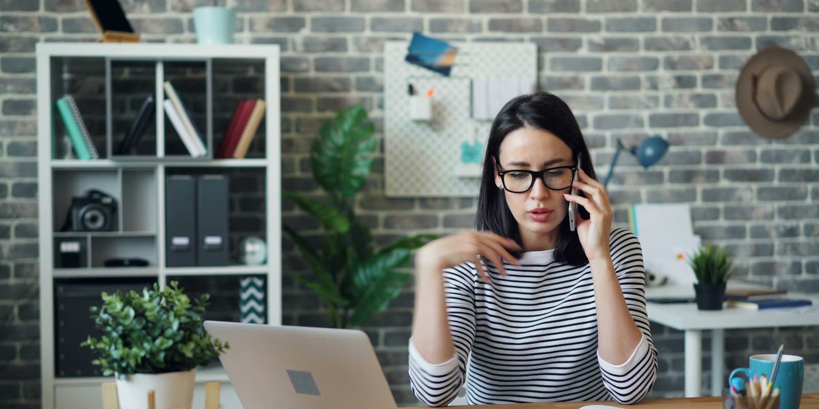 A woman in glasses talking on her phone at a desk with a laptop