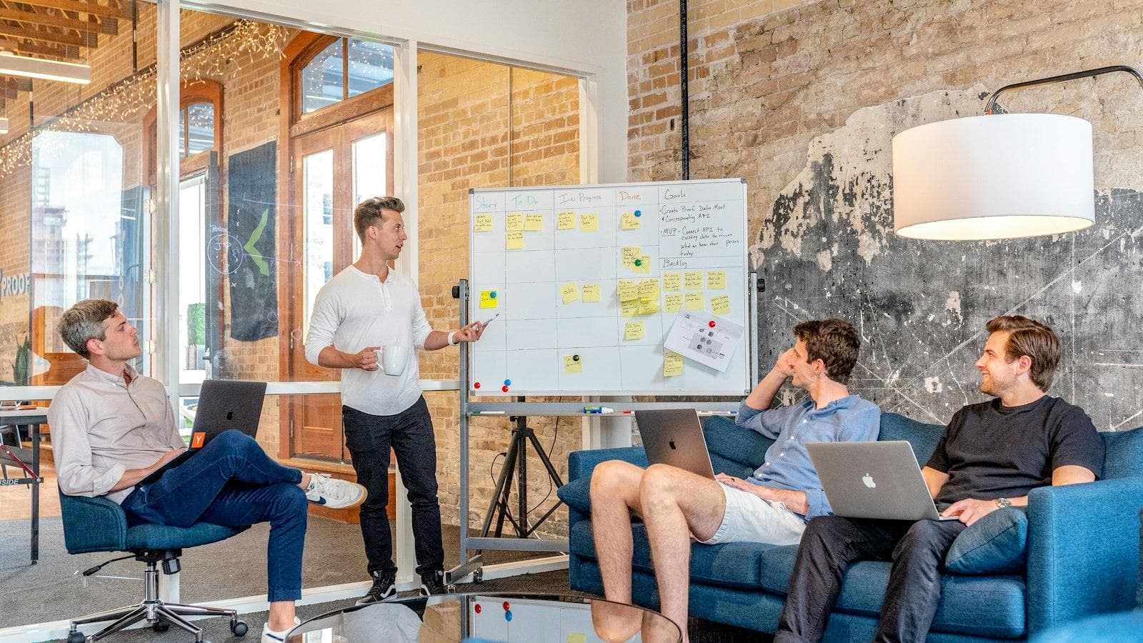 A team lead at a whiteboard of sticky notes talks the plan through with three colleagues on laptops