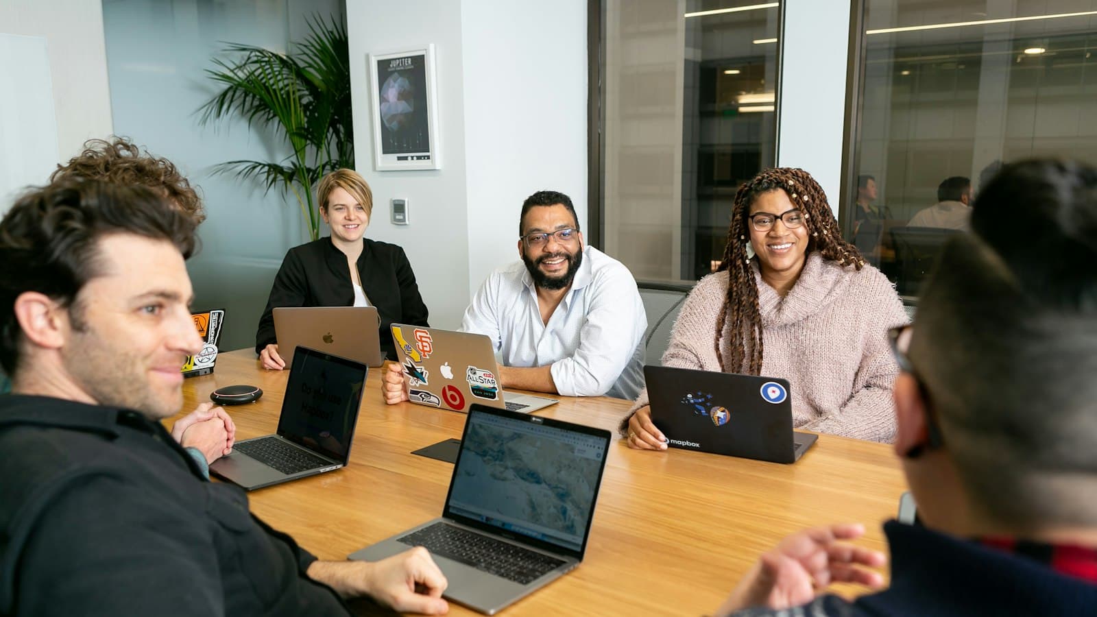 Four colleagues on laptops around a table listening to a team lead