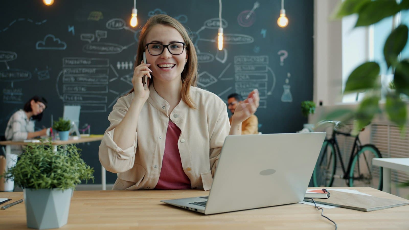 Sales agent smiling while talking on her mobile phone at a laptop in an open office