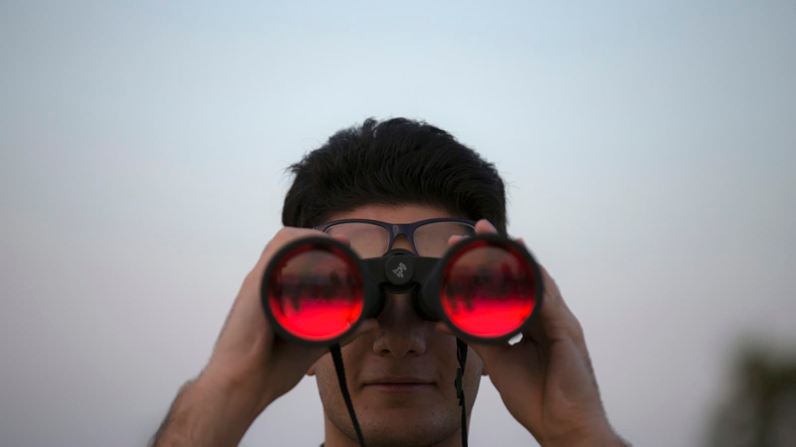 A person looking straight ahead through binoculars against a pale sky