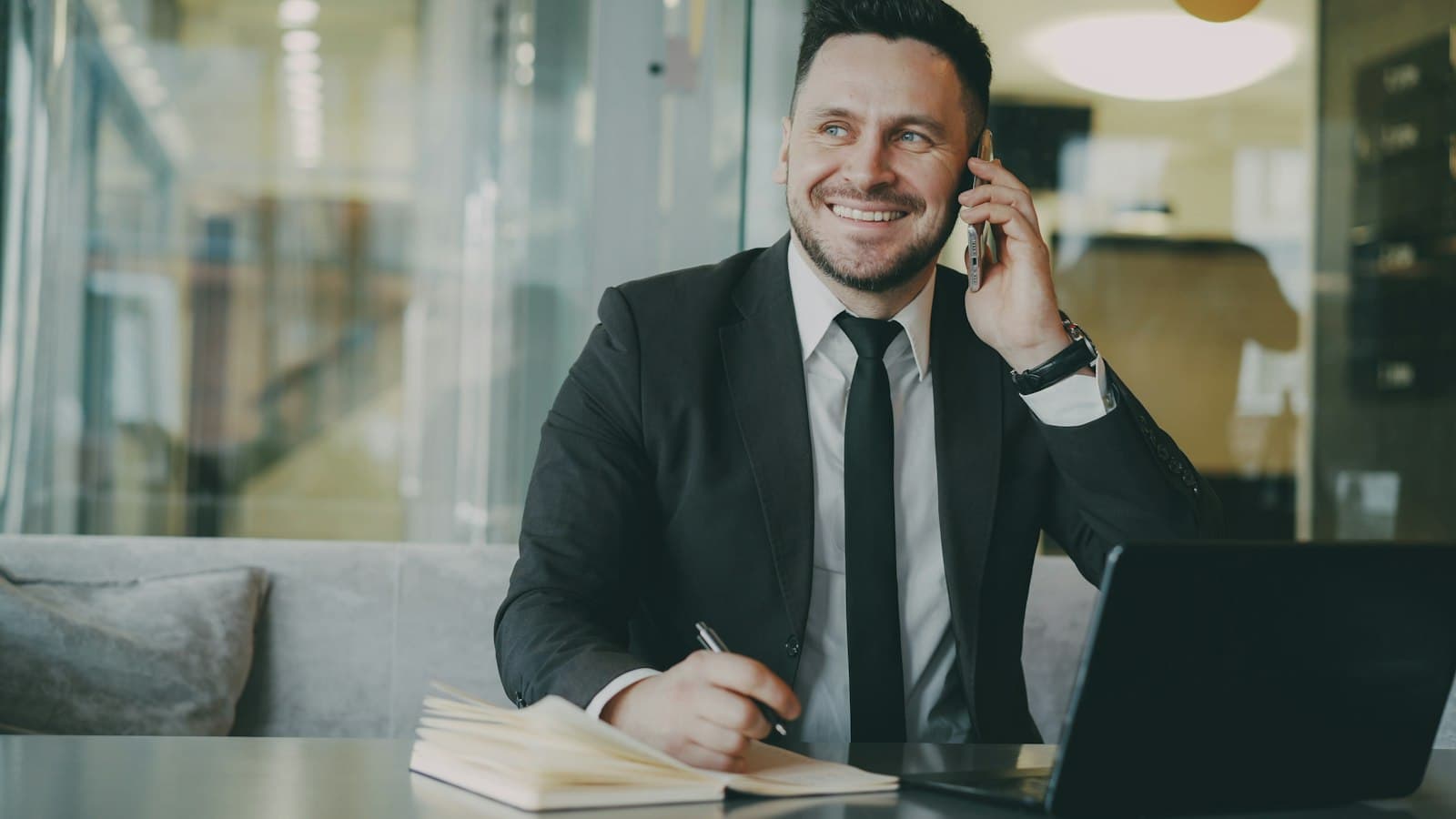 A salesperson in a suit smiles during a phone call, notebook and laptop on the desk