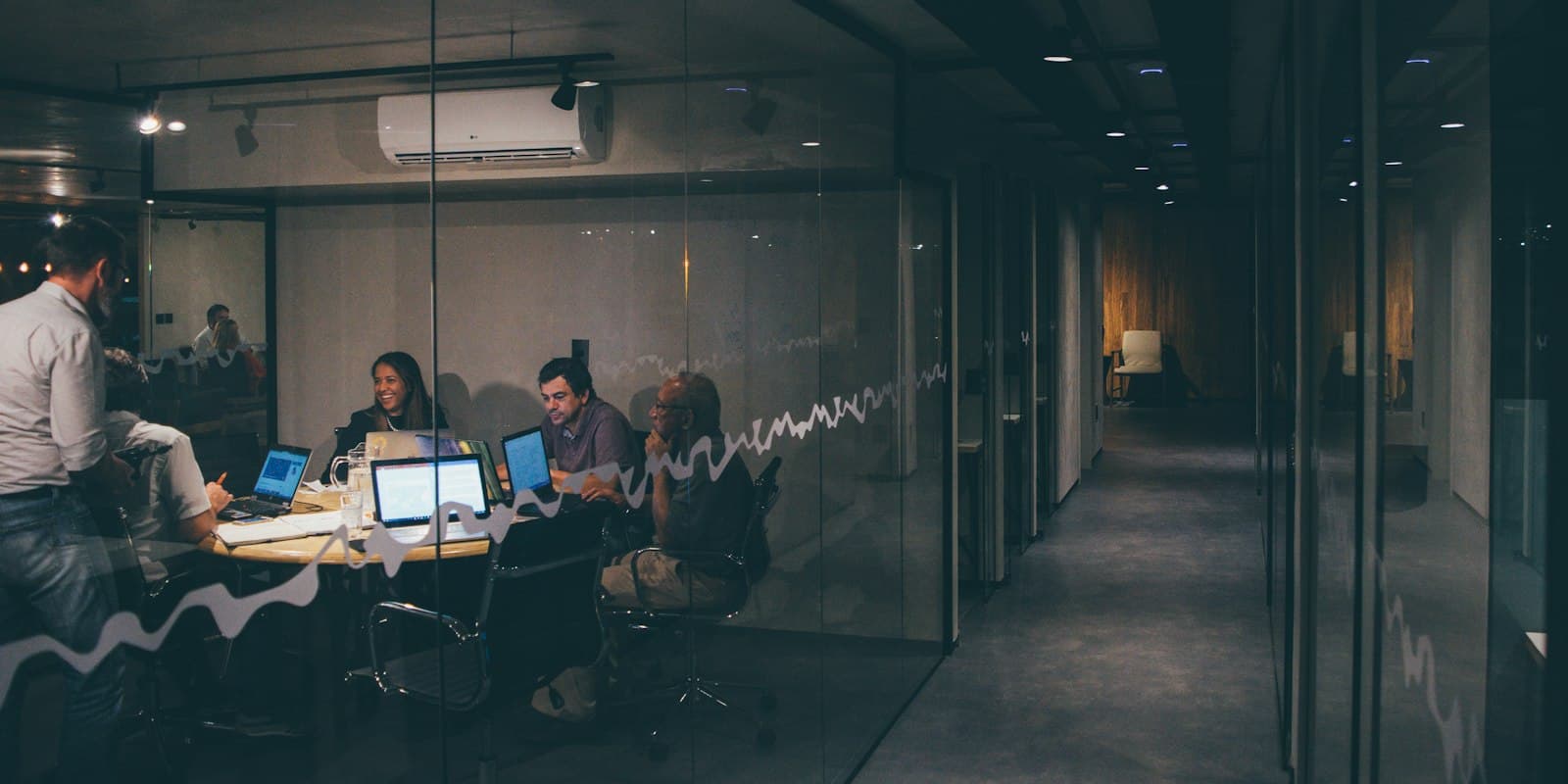 A small team talking around laptops at a meeting table, seen through the glass wall of an office at night