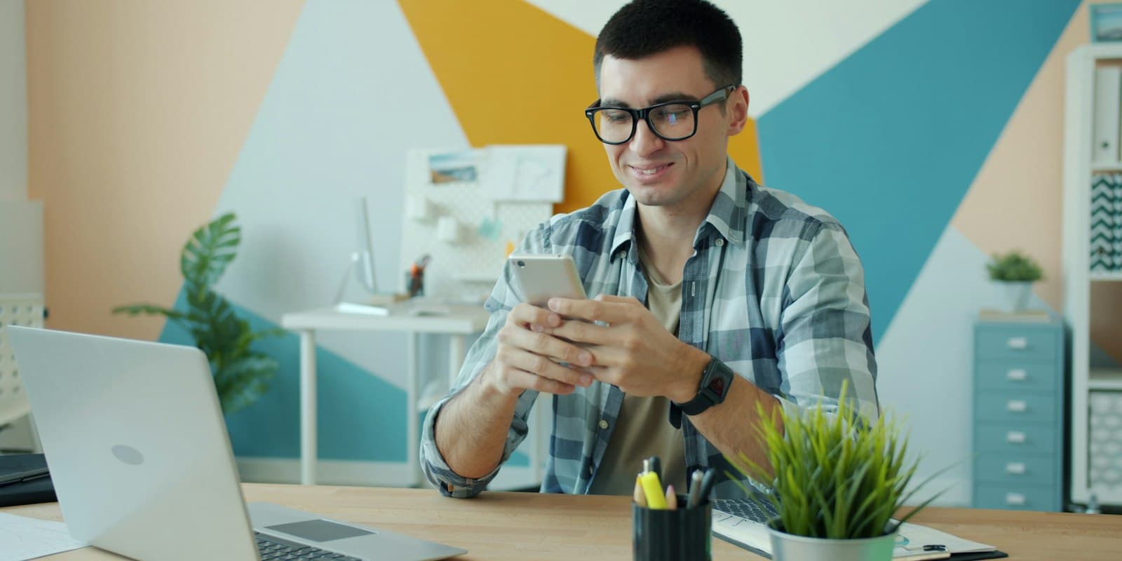 Man in glasses checking his phone at an office desk with a laptop
