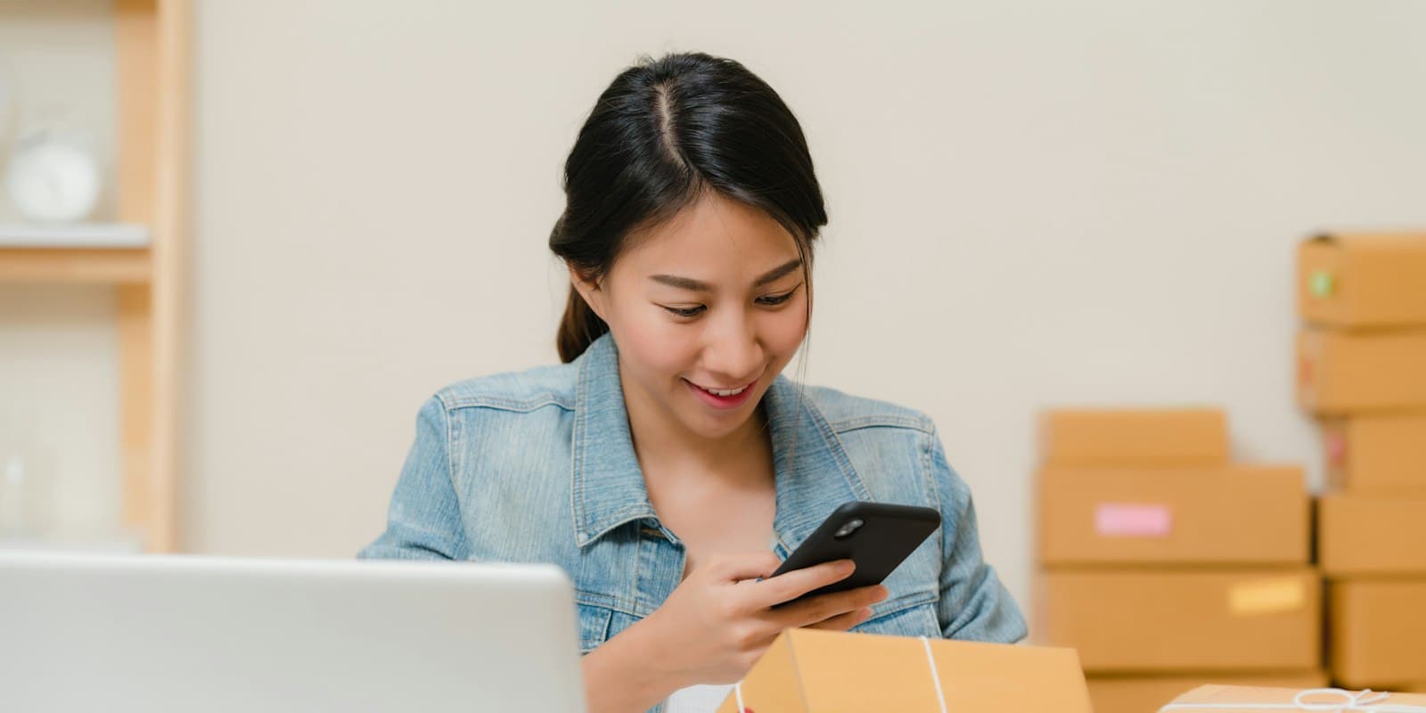 An online seller checking an order on her phone at a desk stacked with parcels