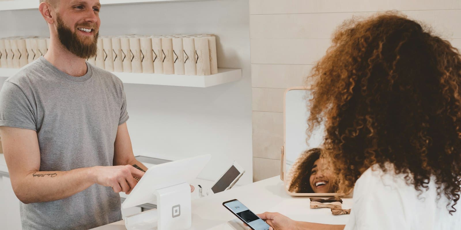 A smiling shopkeeper at his counter while a customer pays with her phone
