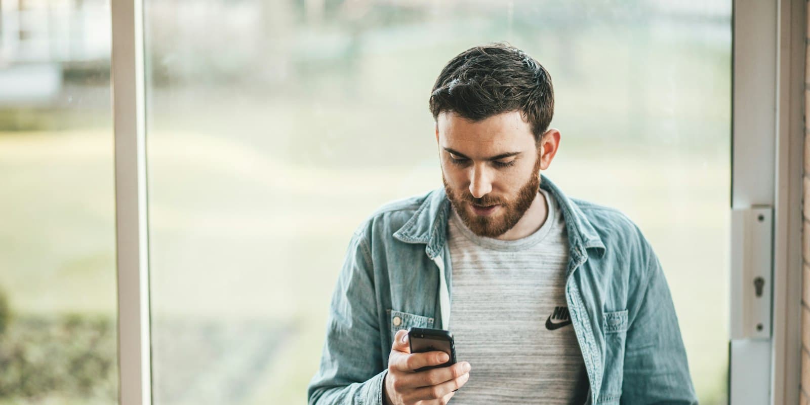 A man by a window looking down at the phone in his hand