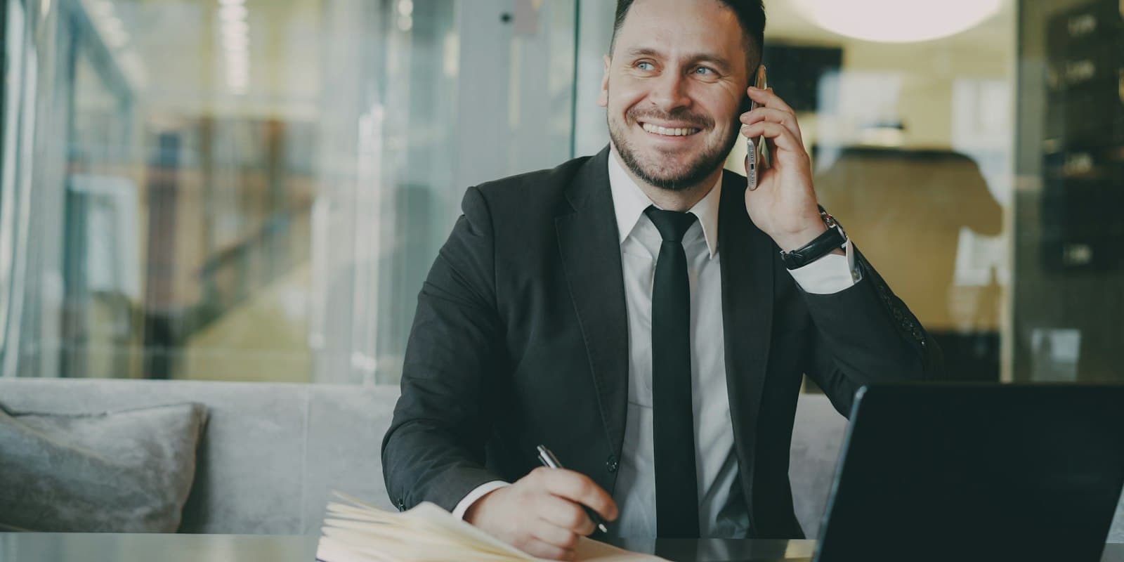 A smiling man in a dark suit talks on his mobile phone at a desk, writing notes beside an open laptop