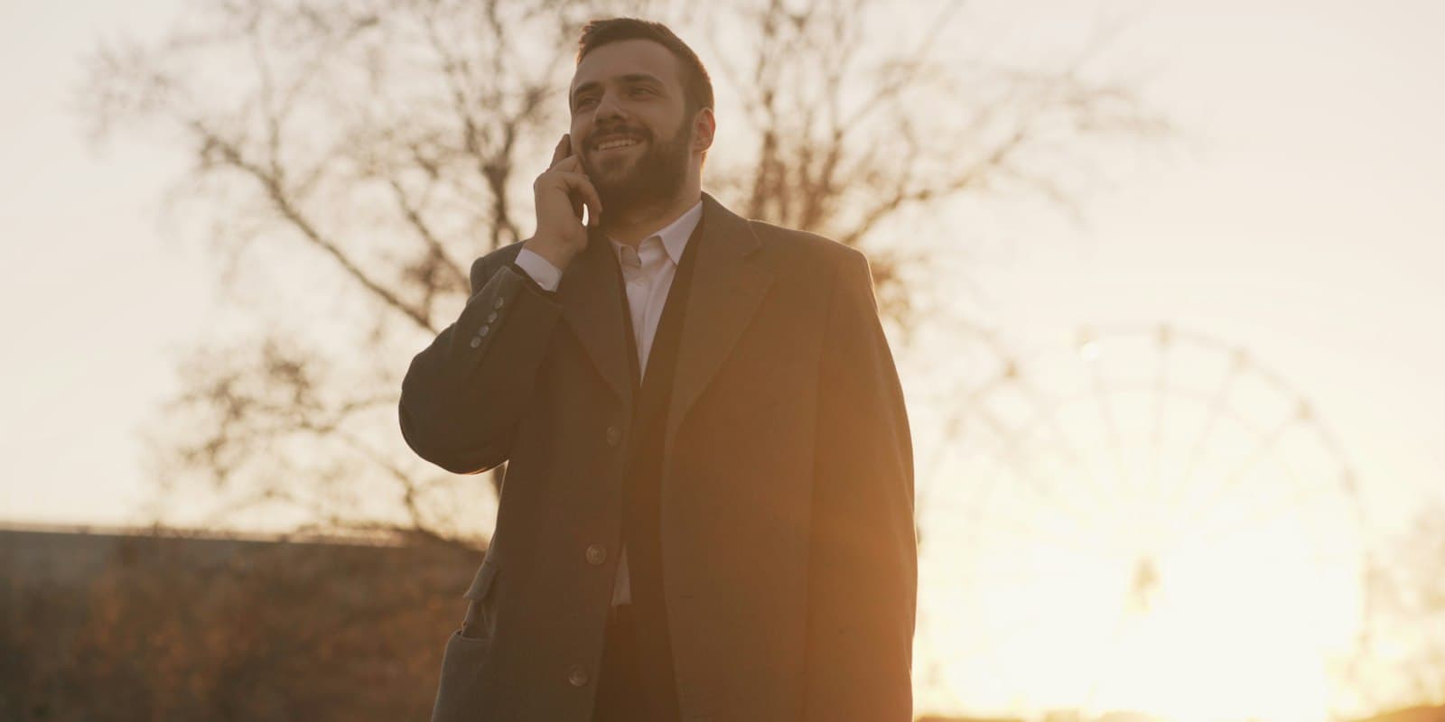 A man in a winter coat smiling as he talks on his mobile phone outdoors at sunset