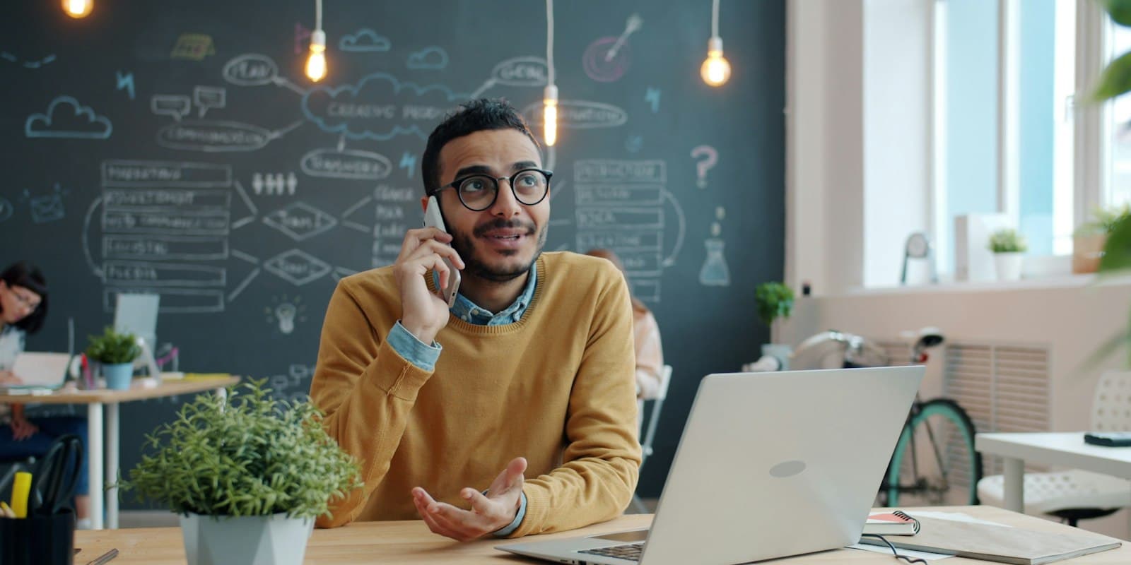 A smiling salesman in a yellow sweater talking on his phone at a desk with a laptop