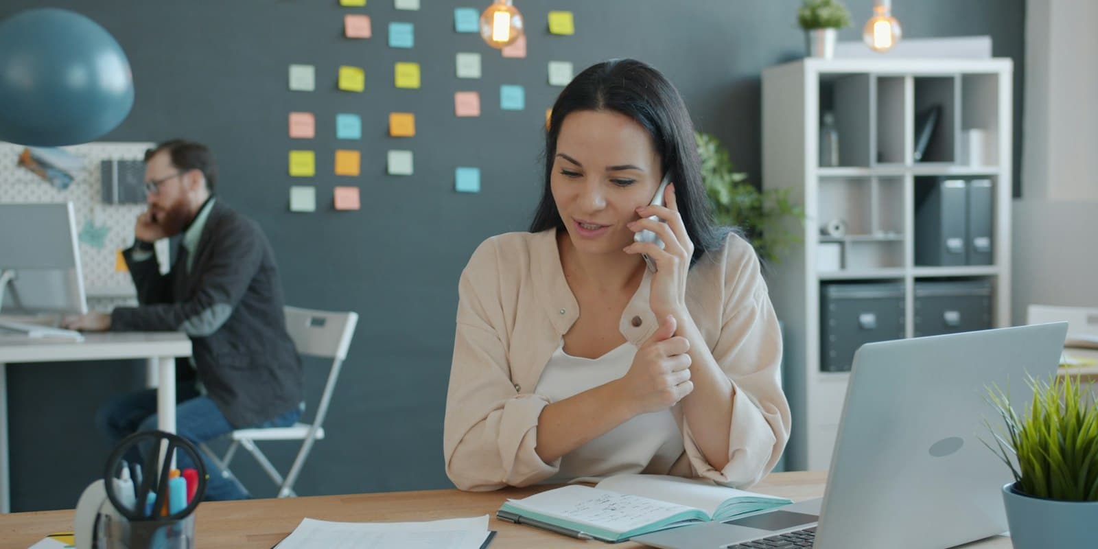 Two colleagues at their desks in a bright office, each talking on a mobile phone