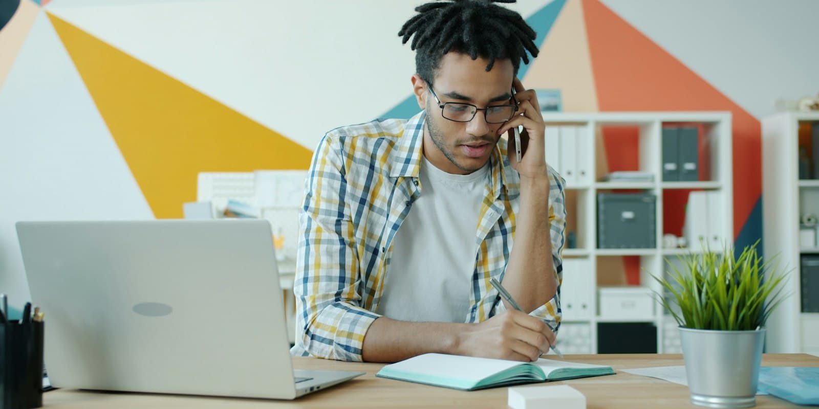 A man on a phone call at his desk, writing notes in a notebook beside his laptop