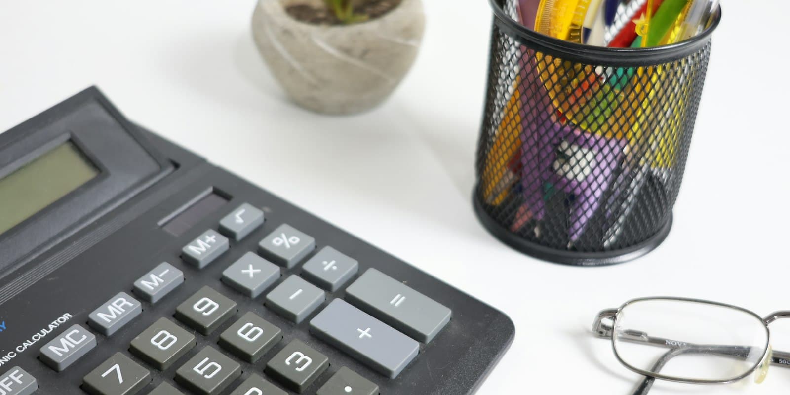 A desk calculator, a pot of pens and a pair of reading glasses on a white desk