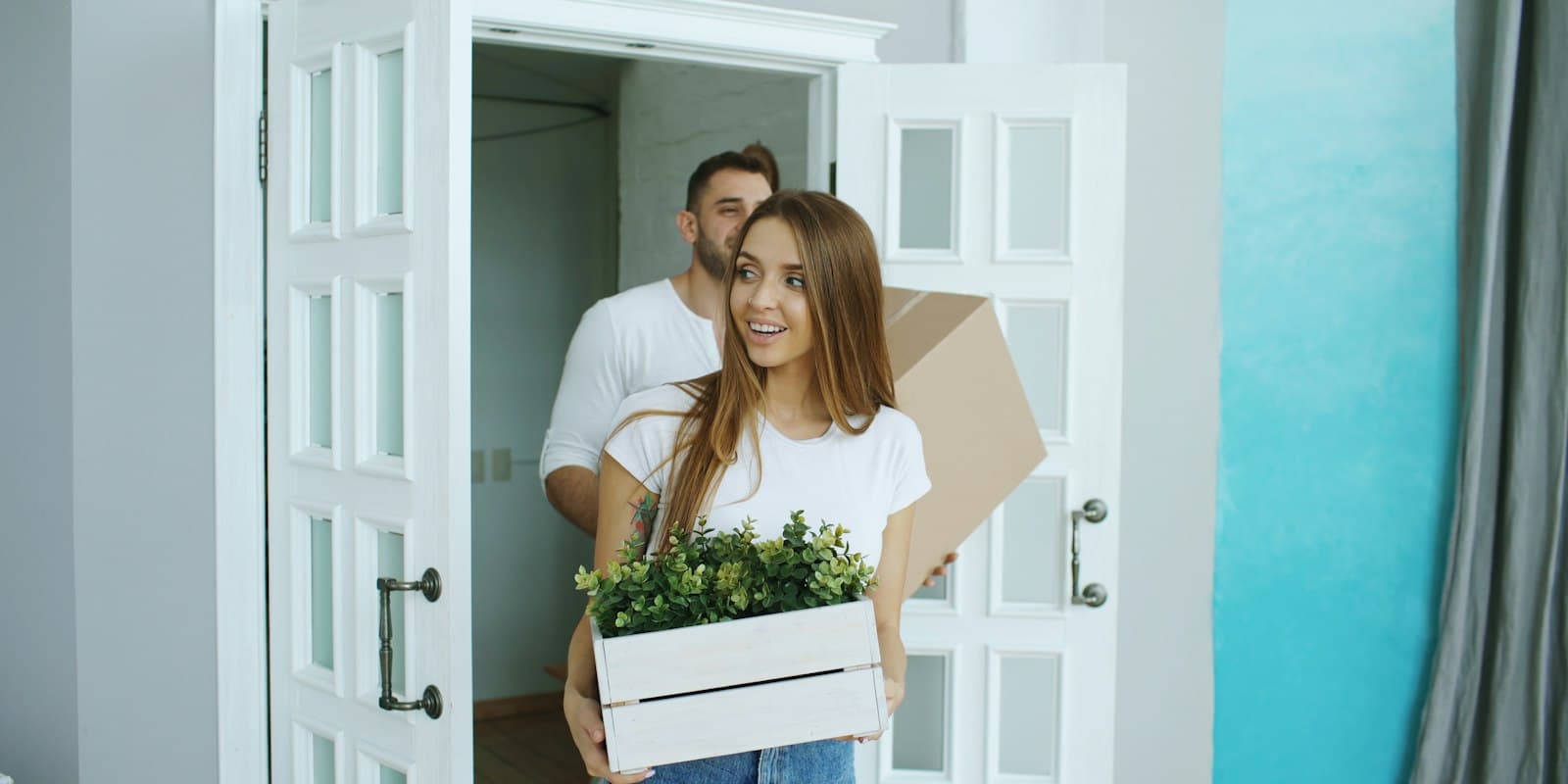 A smiling couple carrying a plant and boxes into a new home