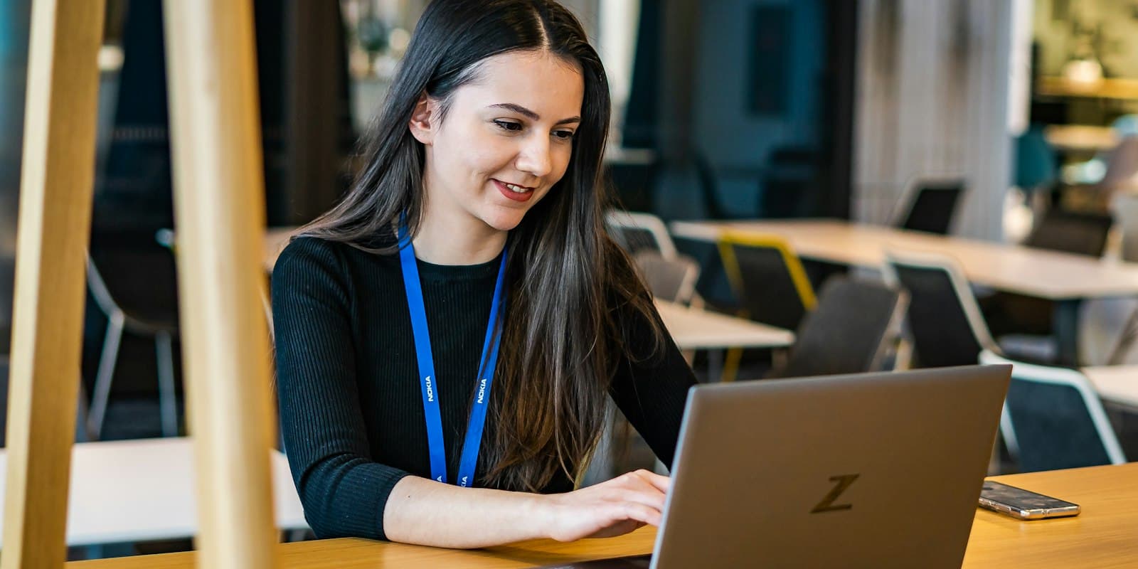 A smiling woman wearing a lanyard, working on a laptop in an office