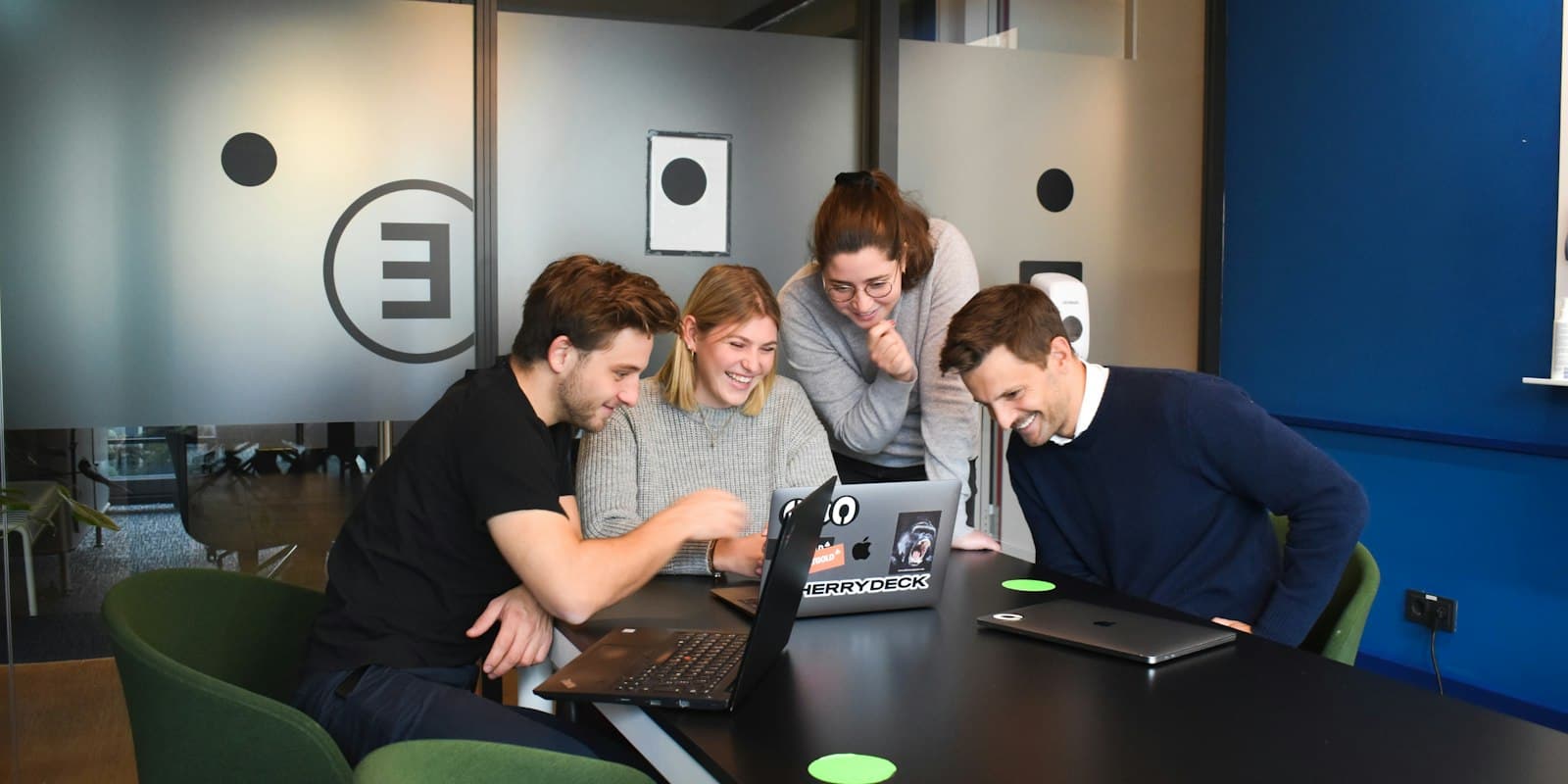 Four colleagues gathered around a laptop in an office, looking at the screen together