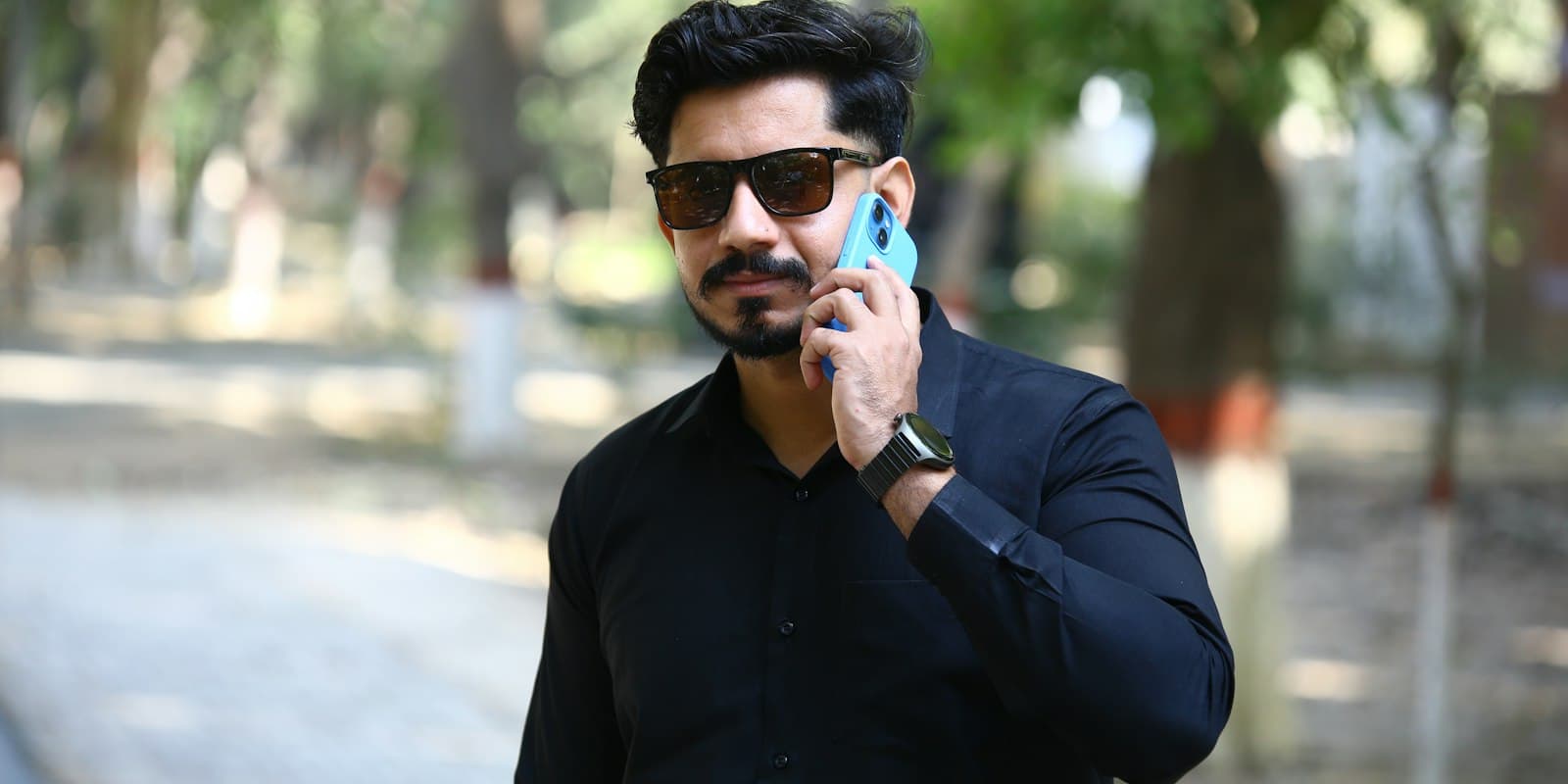 A man in sunglasses and a dark shirt talks on his mobile phone outdoors, with trees behind him